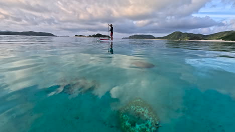 mirror like calm turquoise water reflecting the clouds being illuminated by the rising sun. young sup paddler gently strokes through the panning shot