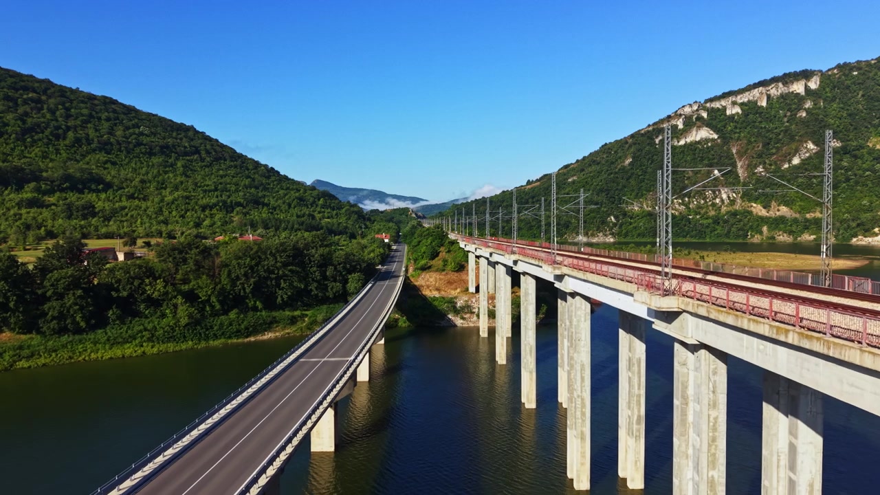 Bridge connects two sides of a river while train tracks run alongside it in a green valley under clear skies