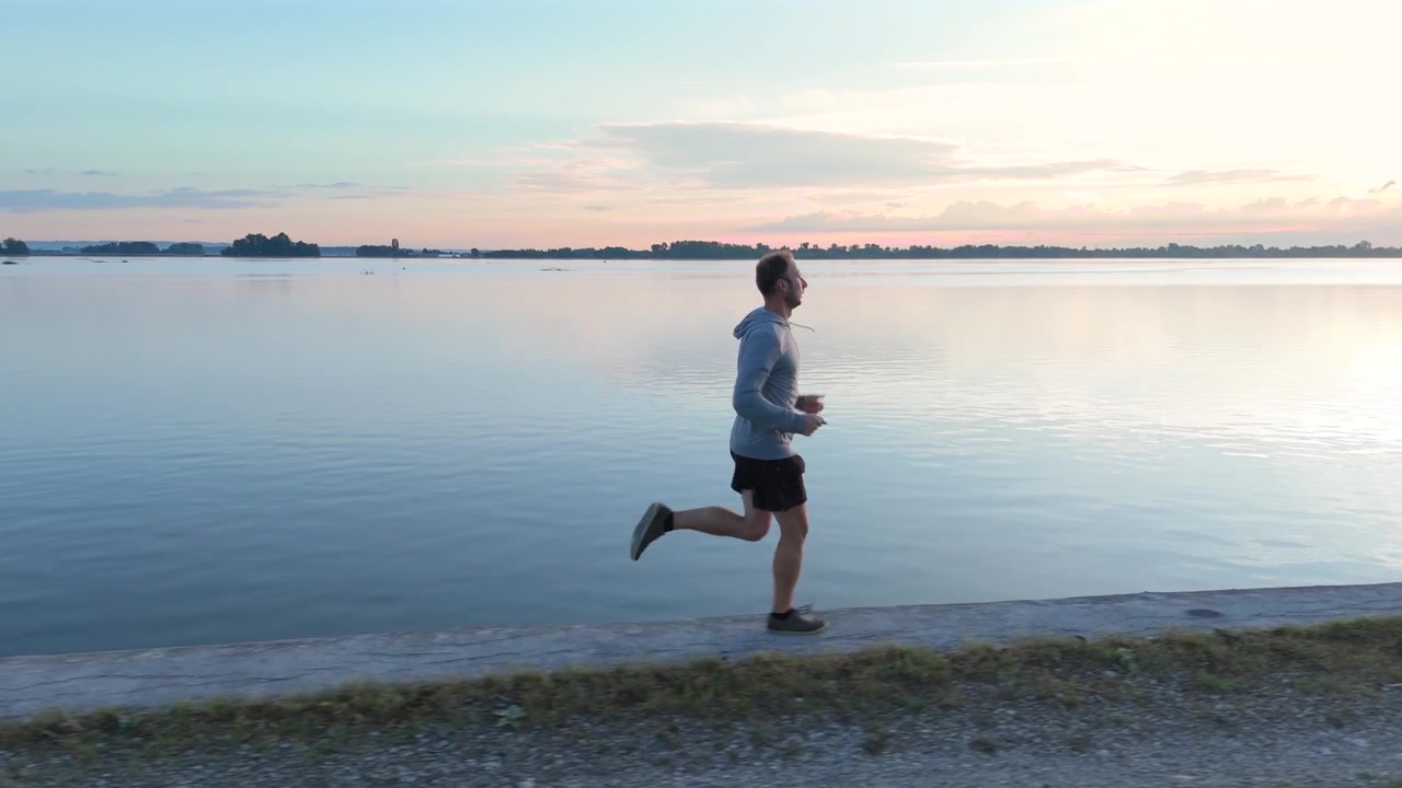 Un drone segue un atleta maschio che corre su un argine fluviale in cemento durante l'ora d'oro. Carrellata cinematografica di un allenamento cardio all'aperto che enfatizza la resistenza e uno stile di vita sano.
