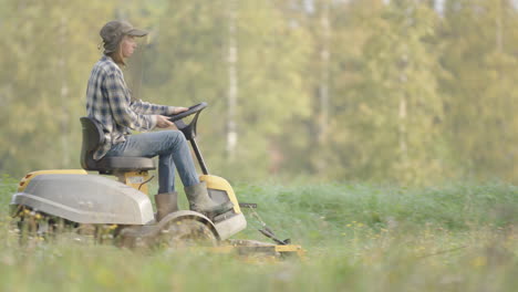 Woman-on-lawn-tractor-mows-grass-wearing-protective-mosquito-headgear-in-summer