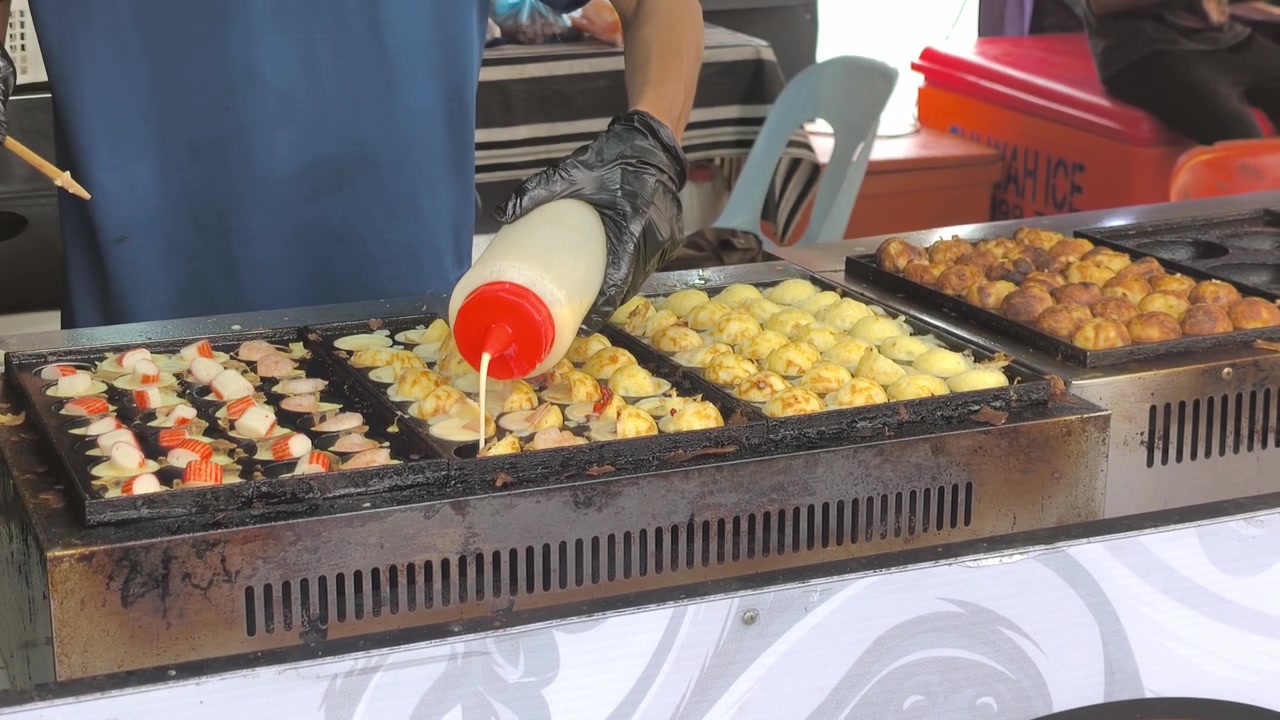 Seorang pedagang kaki lima dengan hati-hati menyiapkan takoyaki segar di atas wajan panas. Bola-bola gurita gurih ini dibalik hingga berwarna cokelat keemasan. Jajanan populer ini menawarkan cita rasa lezat dari kuliner jalanan perkotaan.