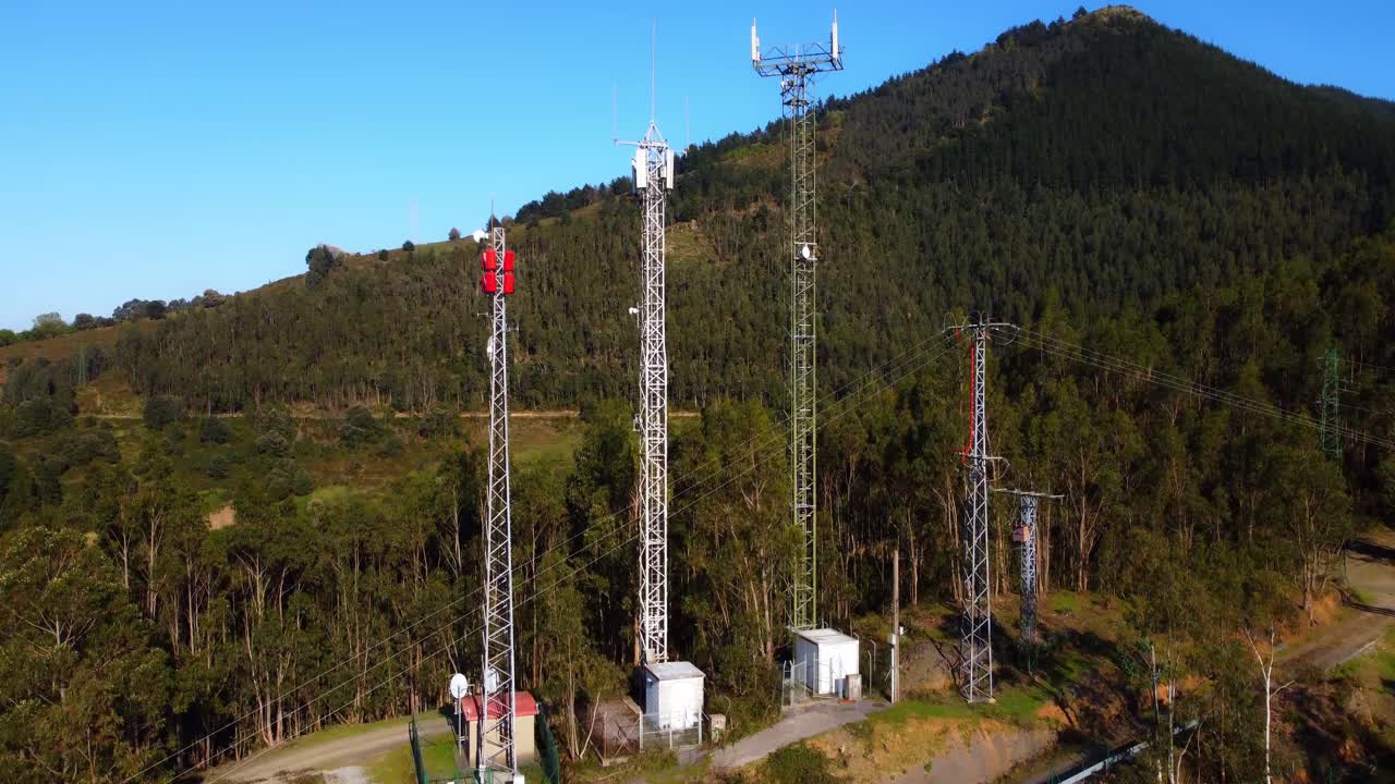 Drone shot rising vertically to reveal three large communication antennas with green hills in the background. Clear summer sky.