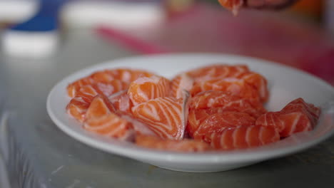 thick slices of raw sake salmon rest on a white ceramic plate placed on a metallic surface, background slightly blurred with hints of red cutting board and utensils, real time, camera close focus