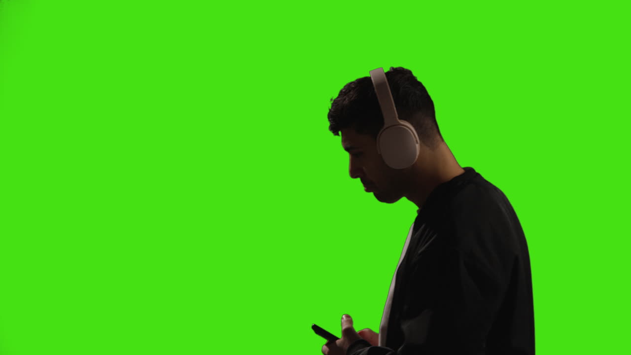 Young Man Wearing Wireless Headphones And Streaming Music Against Green Screen With Low Key Lighting Walking Into Frame
