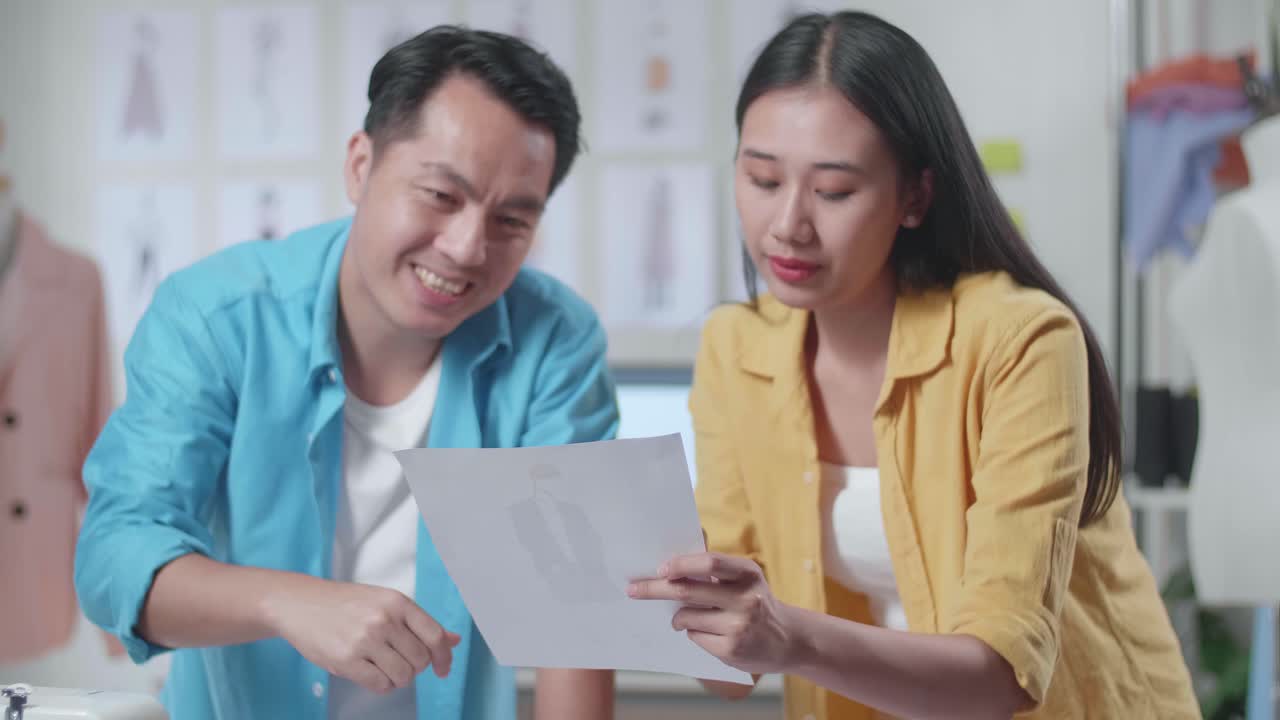 Asian Male And Female Designers With Sewing Machine Discussing About The Clothes Drawing Picture In Hand While Designing Clothes In The Studio