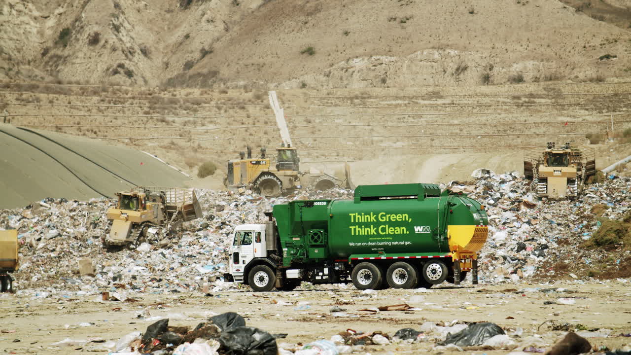 A green Waste Management garbage truck and heavy machinery at a large landfill