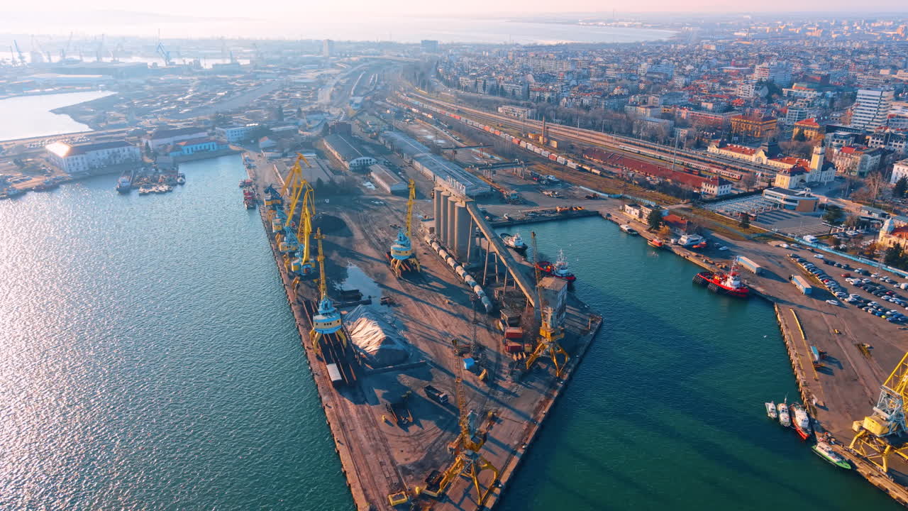 Aerial perspective of port terminal and adjacent city residential area. Elevated view of cargo port infrastructure transitioning into a coastal city with railway tracks and parking