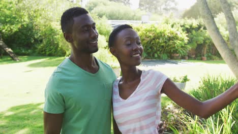 smiling african american couple taking selfie with smartphone embracing in sunny garden