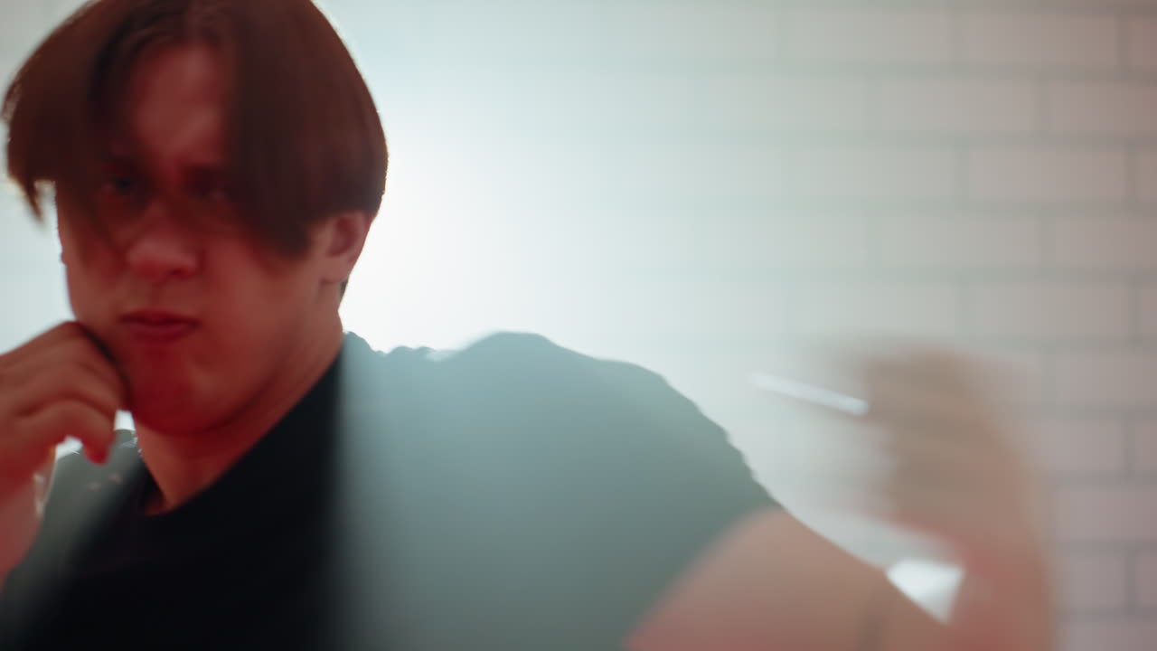 Boxer practicing defensive stance inside gym, wearing black sportswear, positioned against brick wall with bright light in background, showcasing intensity, strength during combat training