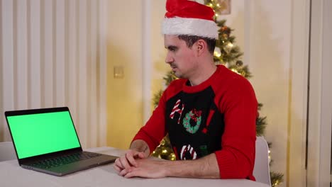 Frightened-young-man-with-Santa-hat-sitting-at-table-next-to-Christmas-tree-and-looking-at-a-laptop-green-screen