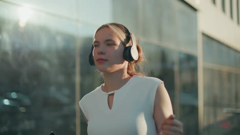 white lady enjoying music with headphones while holding phone in hand, sunlight casting glow on face with hair clip to back, glass building reflecting bright ambiance in urban background