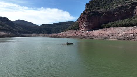 Aerial-view-of-Sau-reservoir-flying-towards-fishing-boat-floating-on-sunny-calm-Ter-river,-Catalonia-mountain-valley