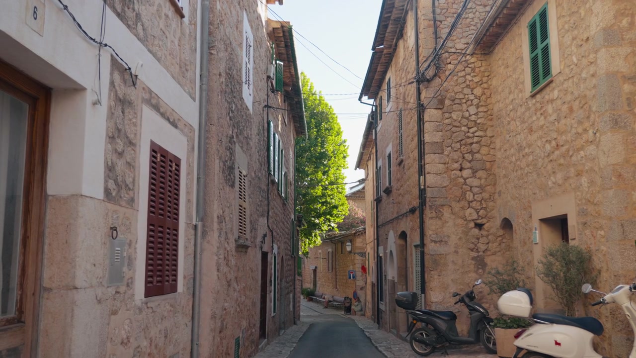 Vista ampia lungo un vicolo stretto di case in pietra color miele con persiane, scooter parcheggiati lungo il muro e un albero verde alla fine. Scena autentica di un villaggio della Tramuntana.
