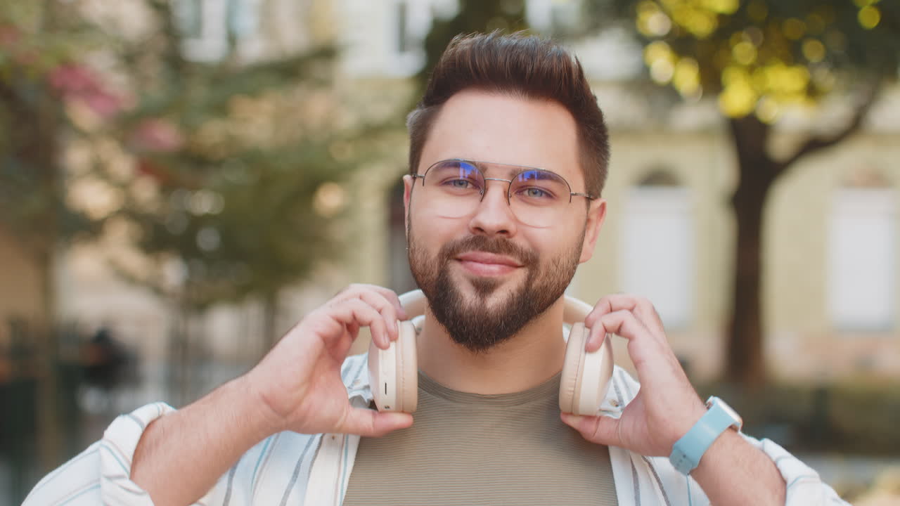 joven caucásico quitándose los auriculares inalámbricos mirando a la cámara y sonriendo en la calle de la ciudad