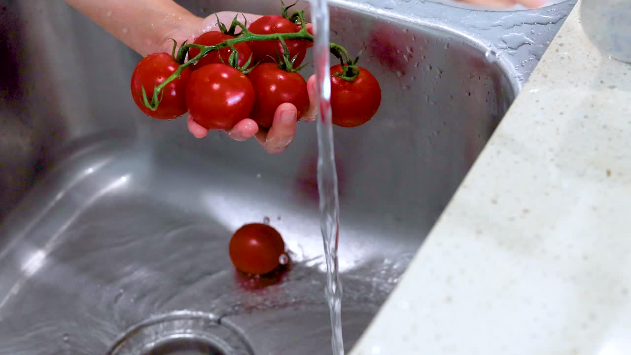 Hands rinse ripe tomatoes under running water in a kitchen sink. Bright lighting highlights the vibrant red color