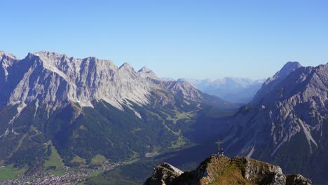 View-of-the-cross-on-the-summit-of-Grubigstein-with-two-hikers-enjoying-the-amazing-views-of-Zugspitze,-Ehrwald-and-Lermoos-in-the-mountains-of-the-alps