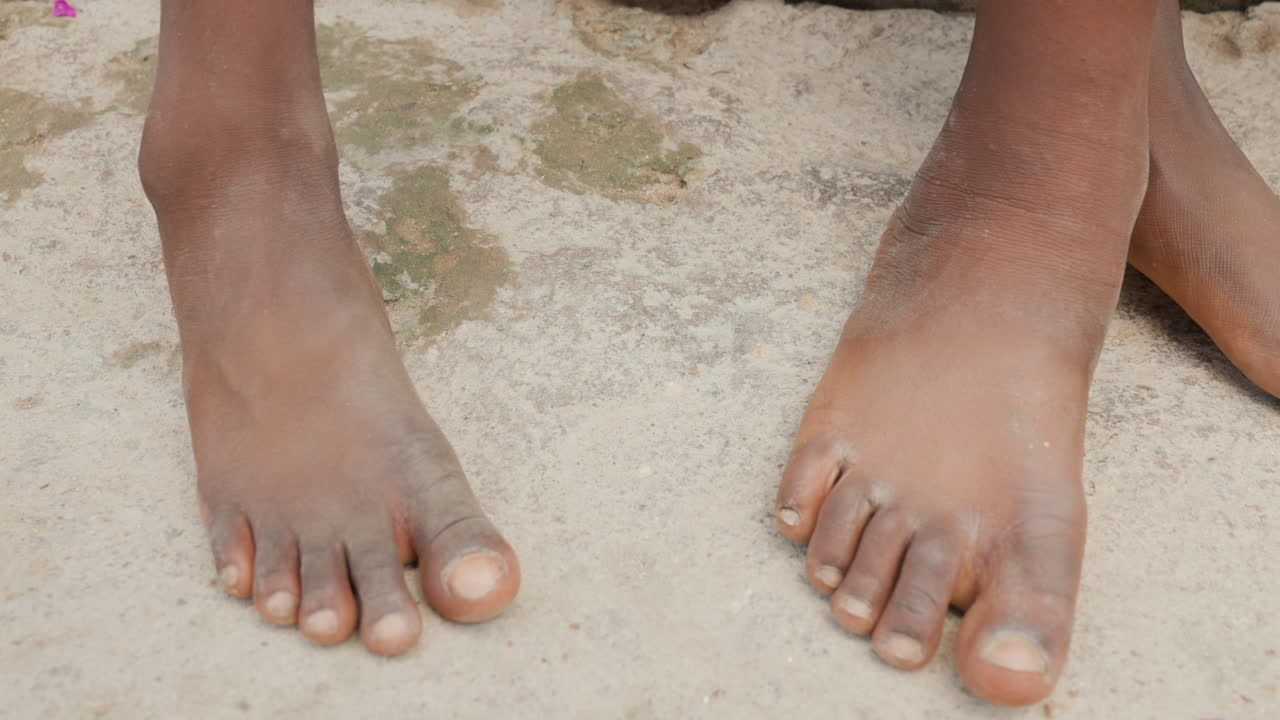 Piedi di bambini di colore su terreno polveroso, primo piano di due paia di dita e piante dei piedi sabbiose, movimento cooperativo che suggerisce una partita di calcio da strada o un gioco di quartiere, con una texture illuminata dal sole e un'atmosfera urbana informale.