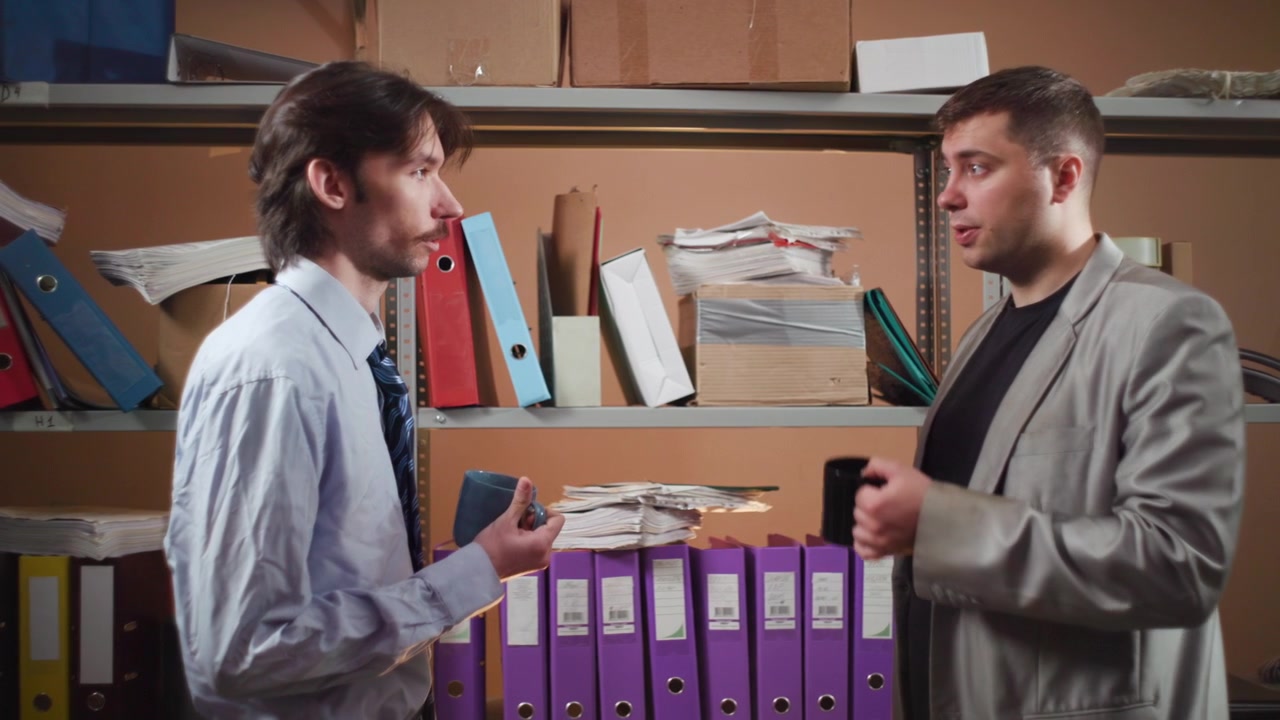 Dos compañeros de trabajo masculinos se miran en un estrecho espacio de almacenamiento de oficina, forrado con estanterías metálicas, archivadores, carpetas y suministros apilados, mientras uno levanta una taza y ambos comparten una seria conversación de negocios.