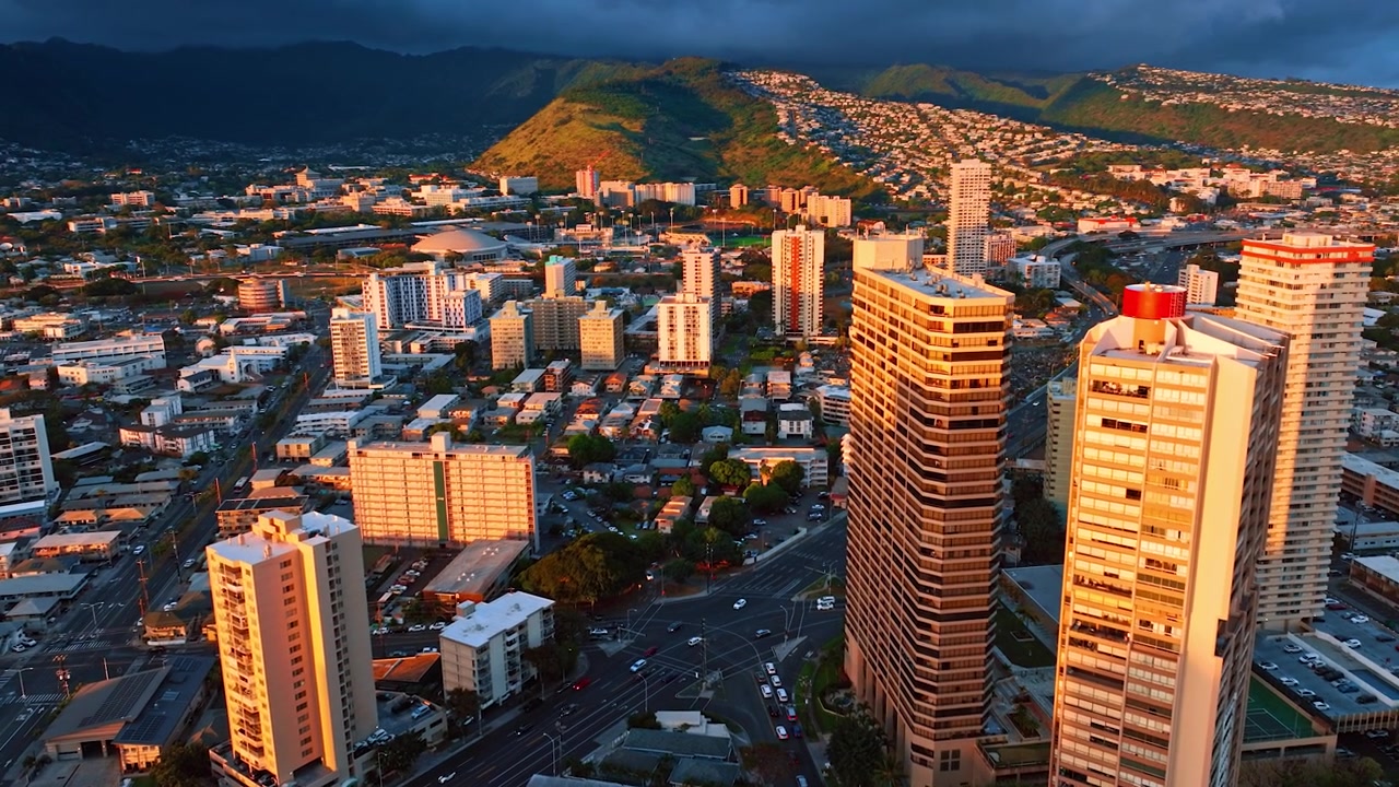 Gli ultimi raggi del sole al tramonto illuminano le cime dei grattacieli di Honolulu e le case sul pendio della montagna. Un cielo scuro e piovoso copre l'orizzonte. Prospettiva aerea