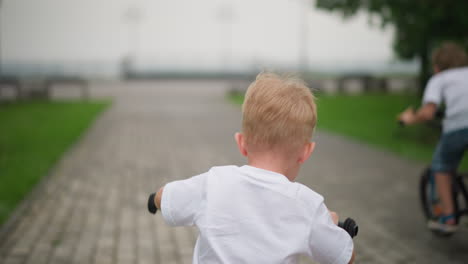 two young children, both wearing white shirts, are seen from the back riding bicycles along an interlocked brick path, with the younger child ahead