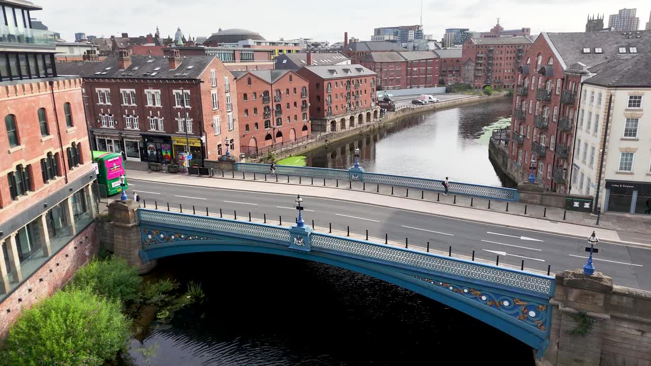 A green double-decker bus travels across a blue bridge in central Leeds, passing Victorian buildings and the River Aire under bright daylight. Aerial perspective, steady camera