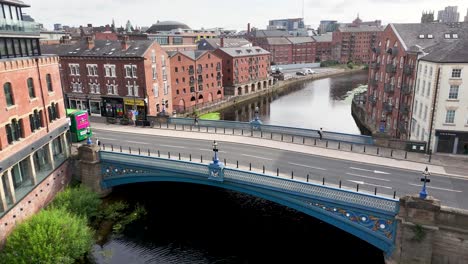 a green double-decker bus travels across a blue bridge in central leeds, passing victorian buildings and the river aire under bright daylight. aerial perspective, steady camera