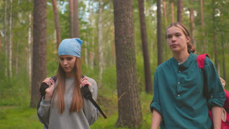 friends walk along forest trail surrounded by lush greenery, one playfully swings her black bag over her shoulder while gazing into distance, while the other remains focused