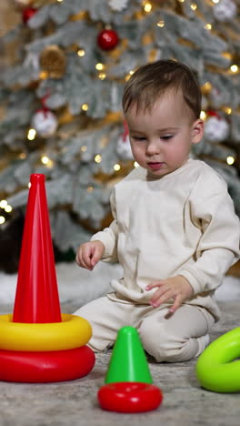 calm caucasian baby builds a toy pyramid sitting on the floor. christmas tree and a black cat under it at backdrop in blur. vertical video