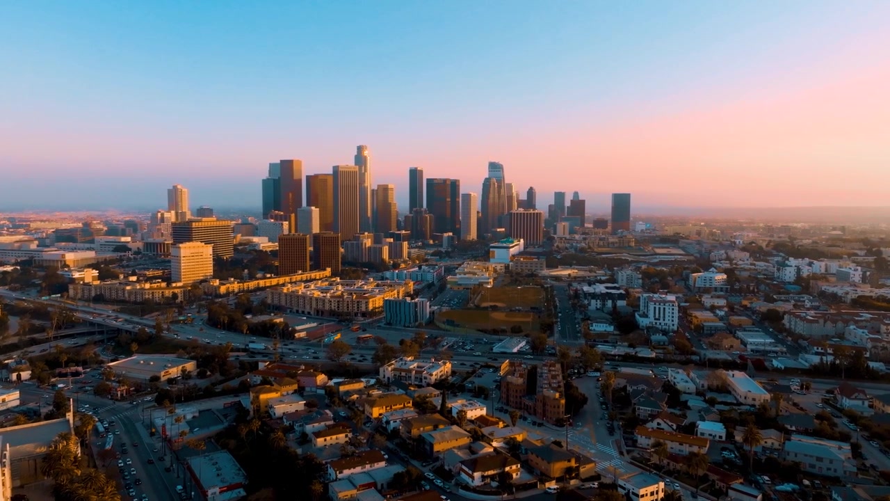 Una vista aerea mostra lo skyline del centro di Los Angeles con grattacieli e traffico congestionato. Palme e infrastrutture urbane sono visibili. L'ora di punta influisce sul flusso dei trasporti