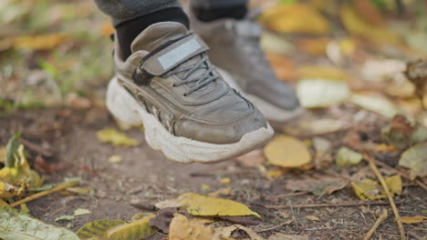 close up of child legs swinging gently above fallen leaves on park ground while scattered foliage drifts in autumn breeze beneath chunky sneakers conveying serene seasonal