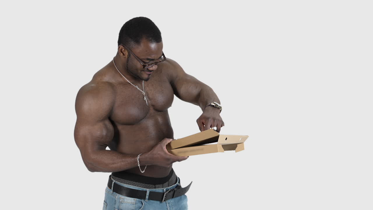 Black skinned sportsman with pizza. Muscled african man in glasses without shirt holding a box and taking out a piece of pizza in studio.