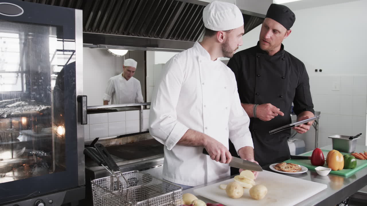 cocinero caucásico enfocado instruyendo a un cocinero aprendiz con una tableta en la cocina, cámara lenta
