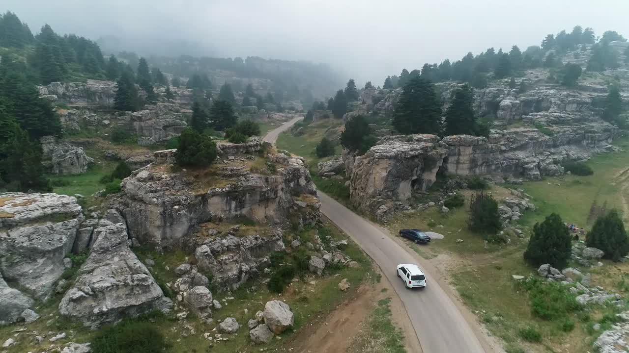 Ripresa aerea di un'auto che attraversa la collina verso una montagna nebbiosa, ricoperta di alberi. Bella e misteriosa natura dell'alta montagna. Drone che si avvicina all'auto seguendola.