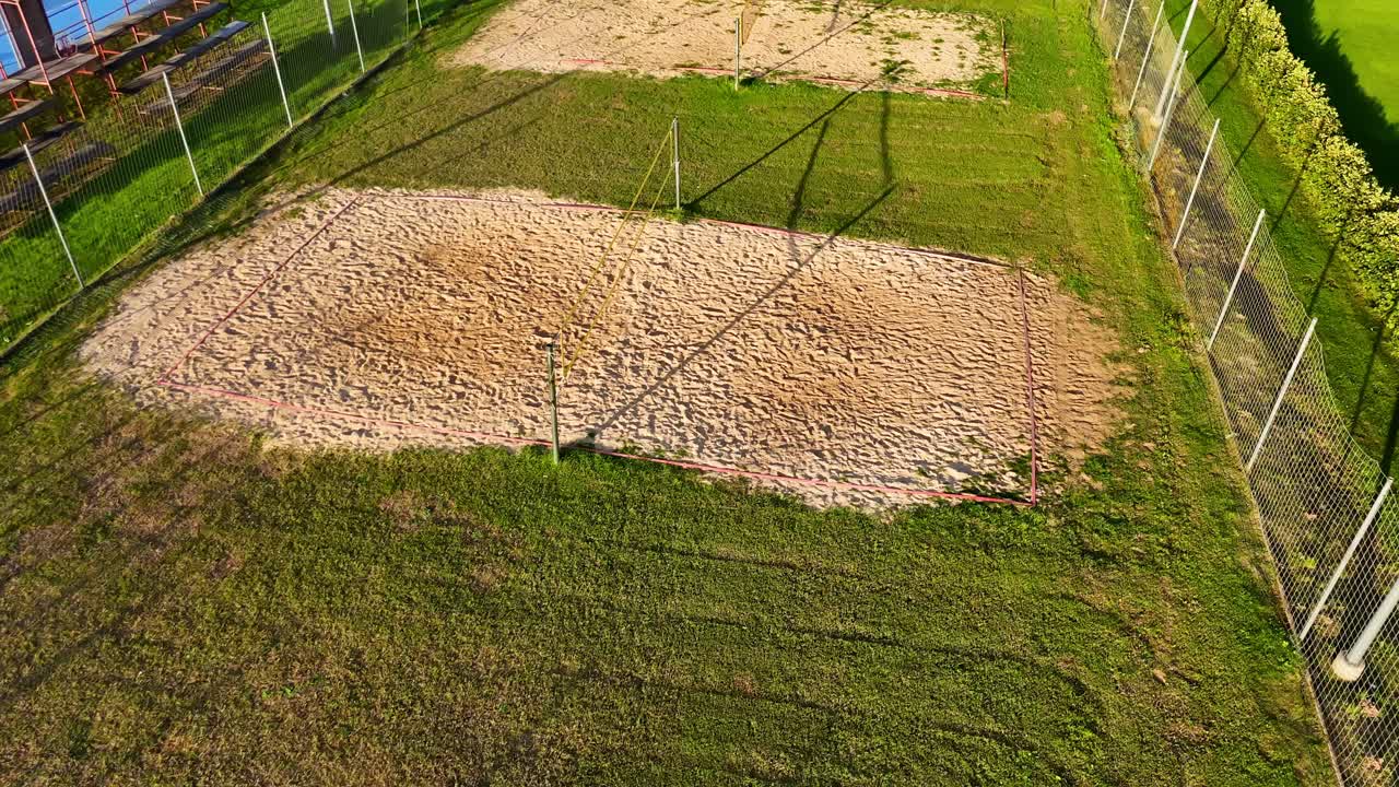Drone view of an empty sand volleyball court surrounded by green grass and fencing, captured in bright sunlight with long shadows and a clean, organized sports layout
