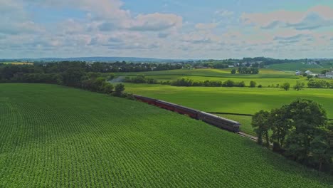 Una-Vista-Aérea-De-Los-Campos-De-Maíz-Y-La-Cosecha-De-Cultivos,-Con-Manchas-De-Color-Con-Un-Tren-De-Pasajeros-Que-Viaja-A-Través-De-él-En-Un-Hermoso-Día-De-Verano