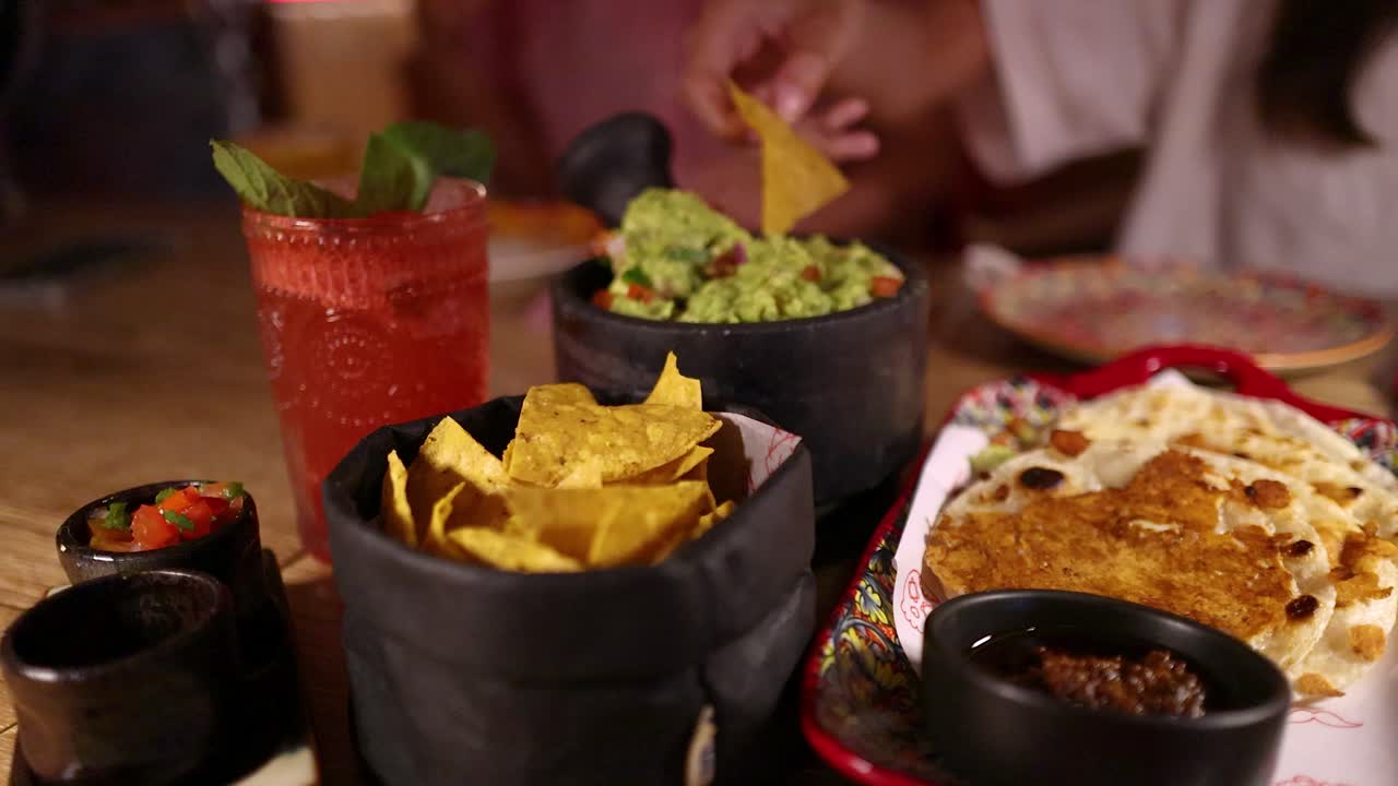 A hand reaches for nacho chips, dips into guacamole beside fresh salsa, enchiladas, and a red cocktail in a warmly lit restaurant setting