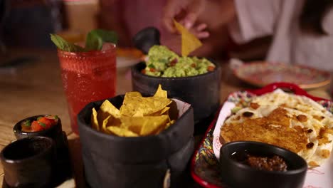 a hand reaches for nacho chips, dips into guacamole beside fresh salsa, enchiladas, and a red cocktail in a warmly lit restaurant setting