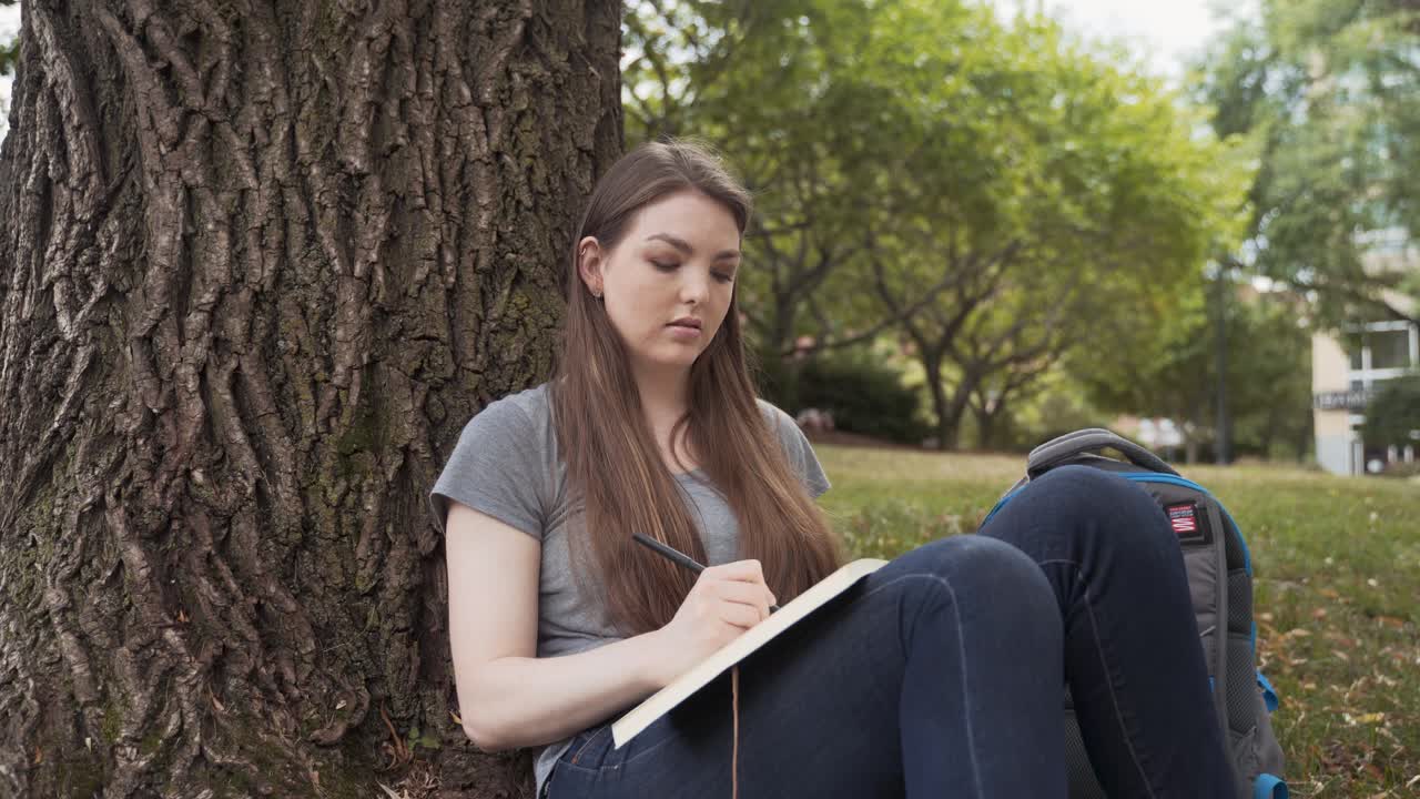 colegiala caucásica escribiendo en un diario sentada bajo un árbol en el parque del campus en un hermoso día