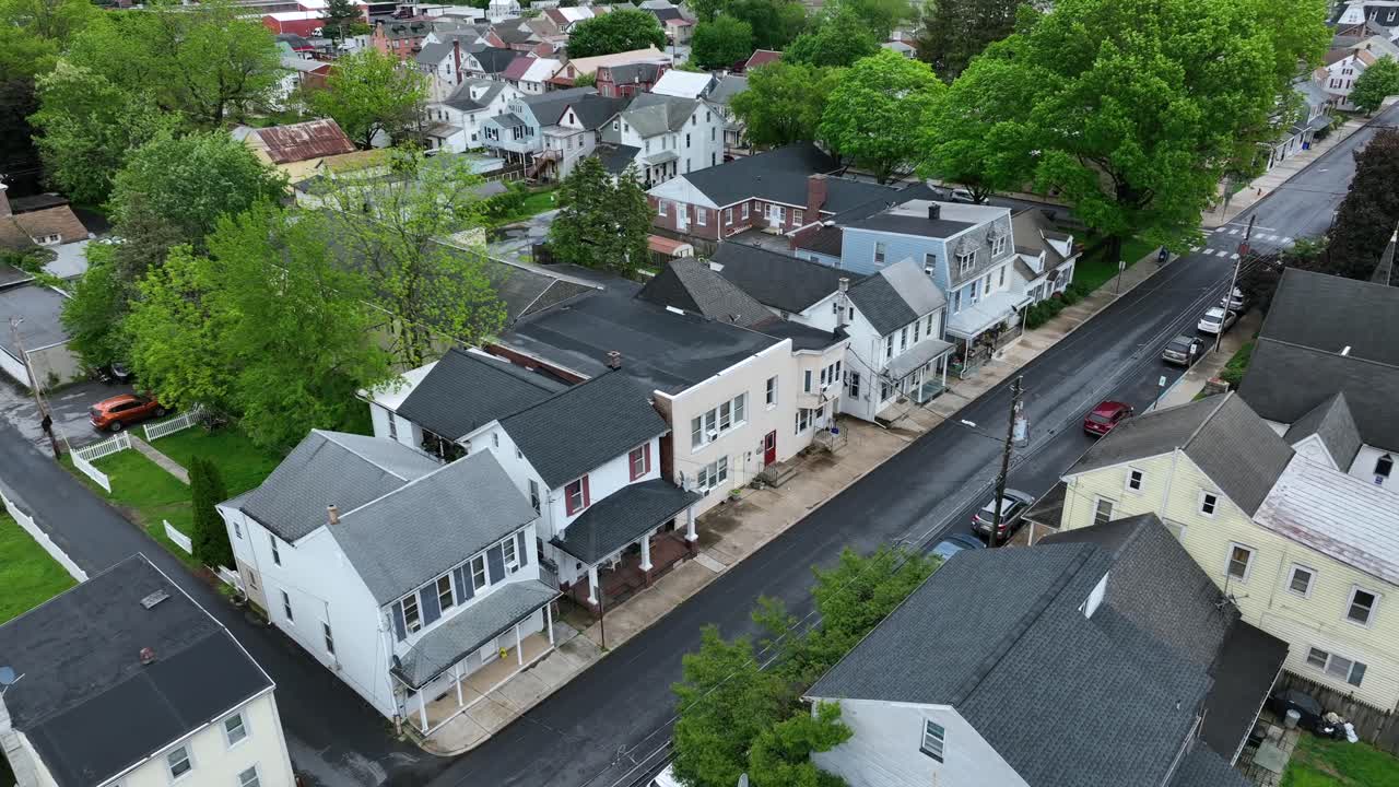 Row of houses with porch along road of american town at cloudy day. Aerial approaching shot. Green trees on cloudy spring day. Top down. Cars on street of quiet housing area in PA, United states.
