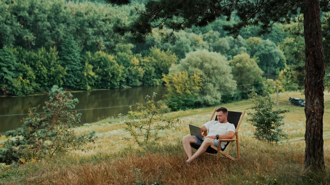 Caucasian freelancer sits in a folding chair surrounded by beautiful nature. Man works on laptop in a comfortable atmosphere remotely from office.