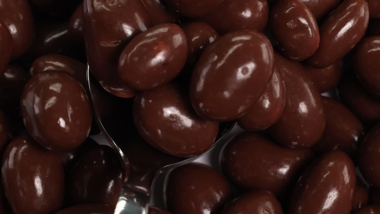 A spoon scoops glossy chocolate-covered peanuts, highlighting texture and shine under soft lighting in a close-up view