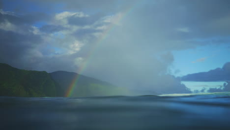 Superficie-Del-Agua-Del-Océano-Mientras-El-Arco-Iris-Cae-En-Exuberantes-Montañas-Verdes-Tropicales-Al-Final-De-La-Ola