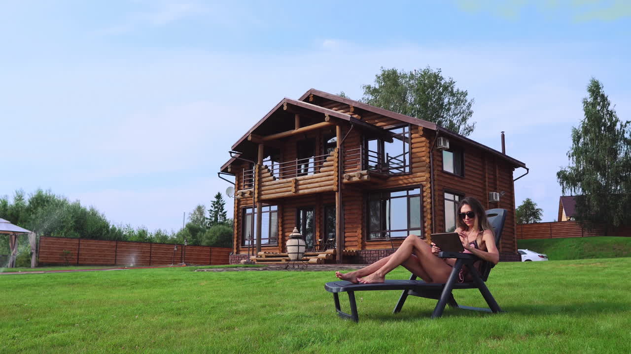 una hermosa morena en traje de baño acostada en una silla larga cerca de su elegante mansión con grandes ventanas sonriendo mientras trabaja en casa de forma remota a través de internet en la esfera empresarial