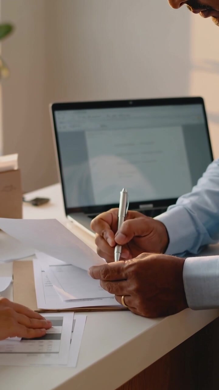 Vertical video: Clipboard shown, man in light blue shirt picking, signing forms at desk with laptop