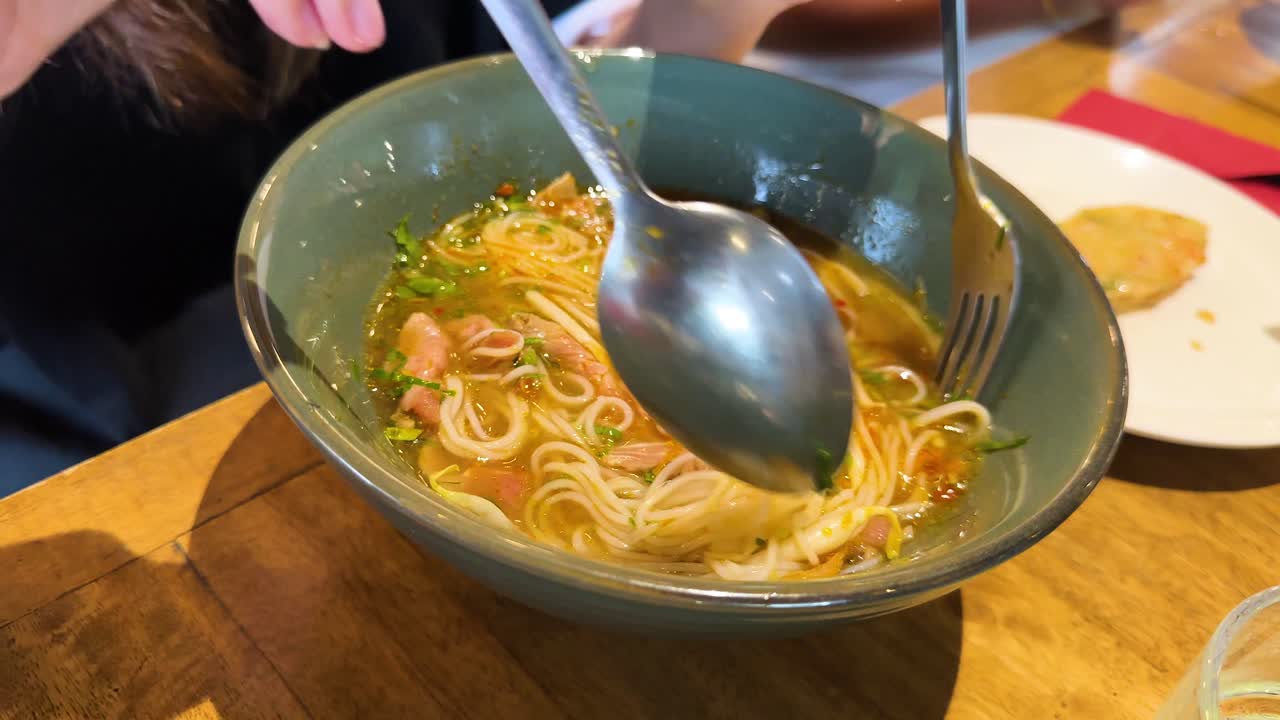 A person stirs a bowl of pho noodles with beef in a Bangkok restaurant. Warm lighting enhances the inviting atmosphere