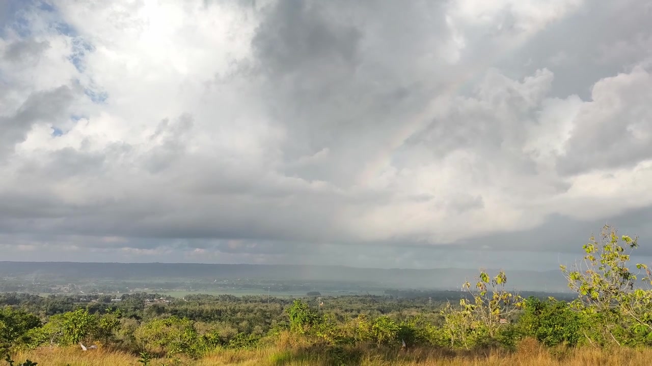 Pelangi Samar di Atas Lembah Hijau Subur di Bawah Langit Berawan