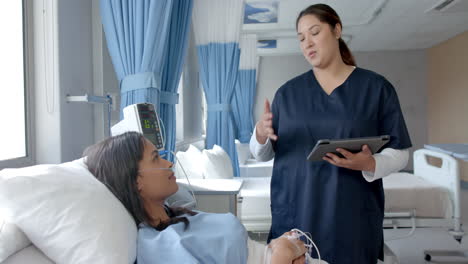 diverse female patient in hospital bed and female doctor using tablet talking, slow motion