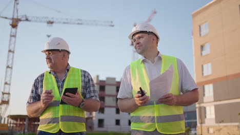 Portrait-of-two-builders-standing-at-building-site.-Two-builders-with-drawings-standing-on-the-background-of-buildings-under-construction-in-helmets-and-vests