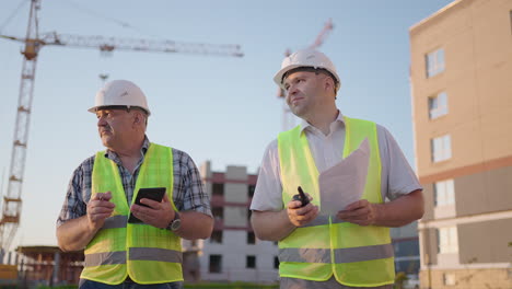 portrait of two builders standing at building site. two builders with drawings standing on the background of buildings under construction in helmets and vests