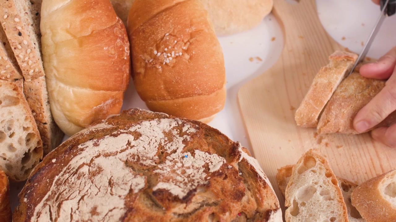 Close-up shot of hands slicing crusty bread on a wooden board among assorted bakery items.
